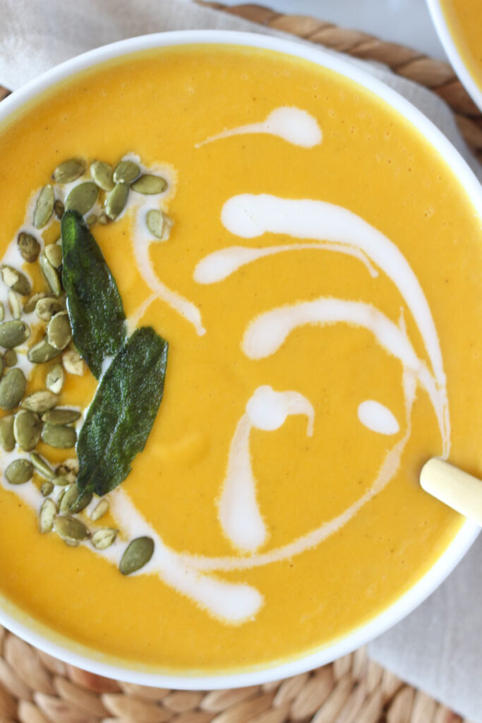 close up of roasted butternut squash apple soup in white bowl topped with pumpkin seeds, fresh sage, and a swirl of coconut milk