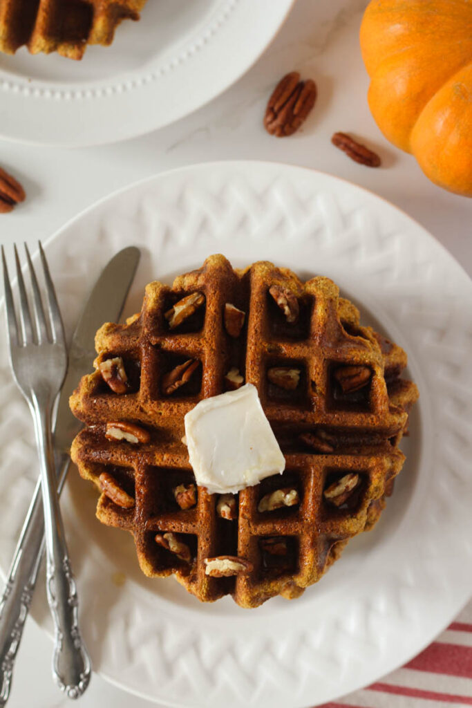 Gluten free pumpkin waffles on white plate with fork and knife topped with chopped pecans, butter, and maple syrup