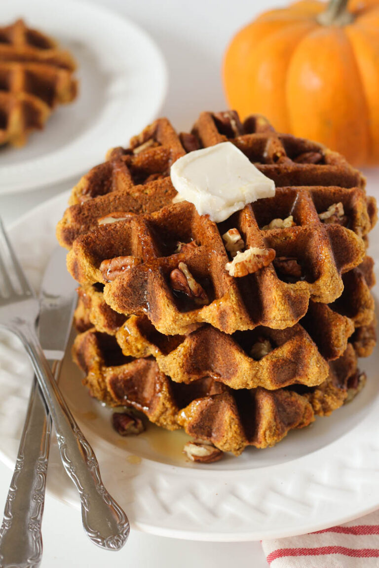 Stack of three gluten free pumpkin waffles on white plate with fork and knife topped with butter, maple syrup, and chopped pecans