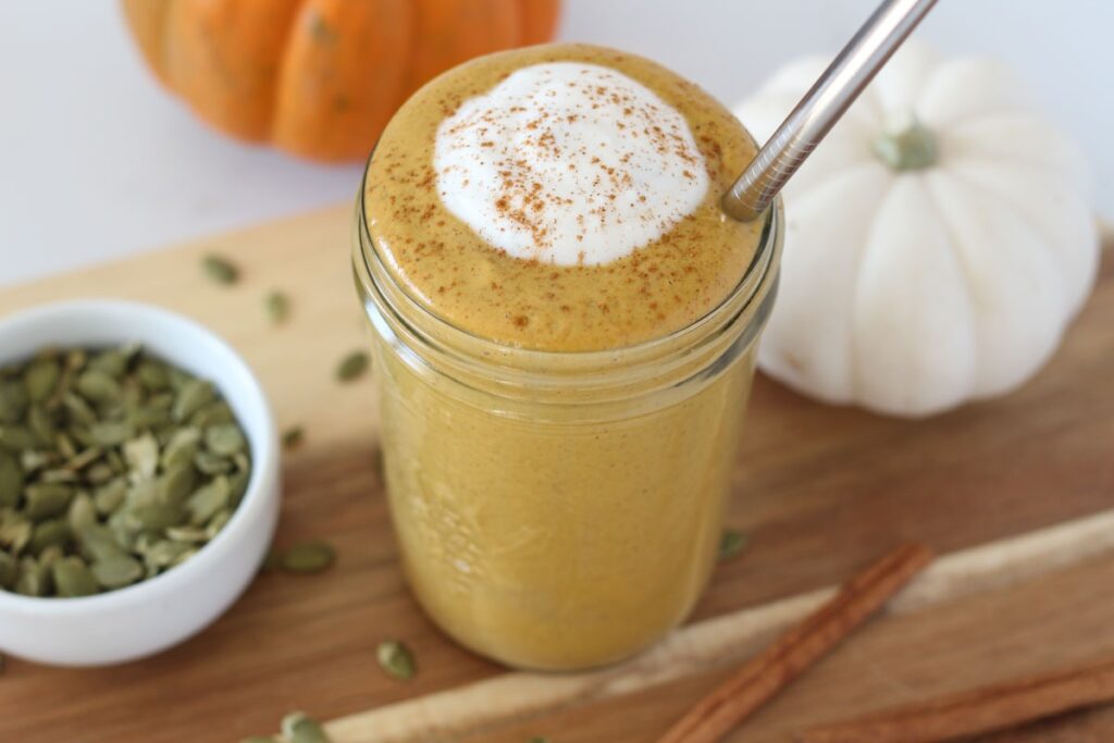 pumpkin spice protein smoothie in mason jar garnished with coconut cream and sprinkle of cinnamon with stainless steel straw on wooden board