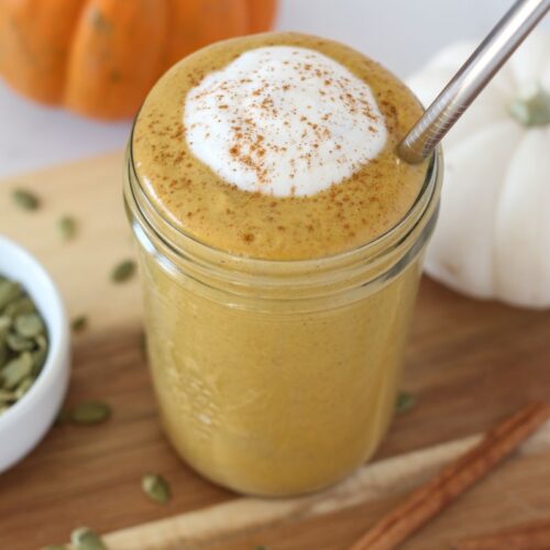 pumpkin protein smoothie in mason jar toped with cinnamon and coconut cream with stainless steel straw sitting on wooden board